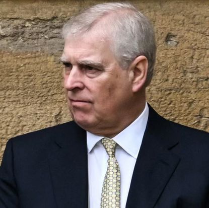 Prince Andrew wearing a suit and tie and looking angry standing in front of a wall