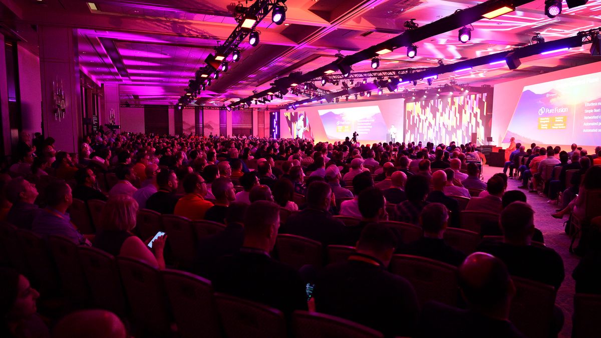 The crowd at Pure Storage&#039;s annual conference Pure Accelerate 2024, watching the opening keynote in Las Vegas. Decorative: The crowd is lit in orange and pink lighting.