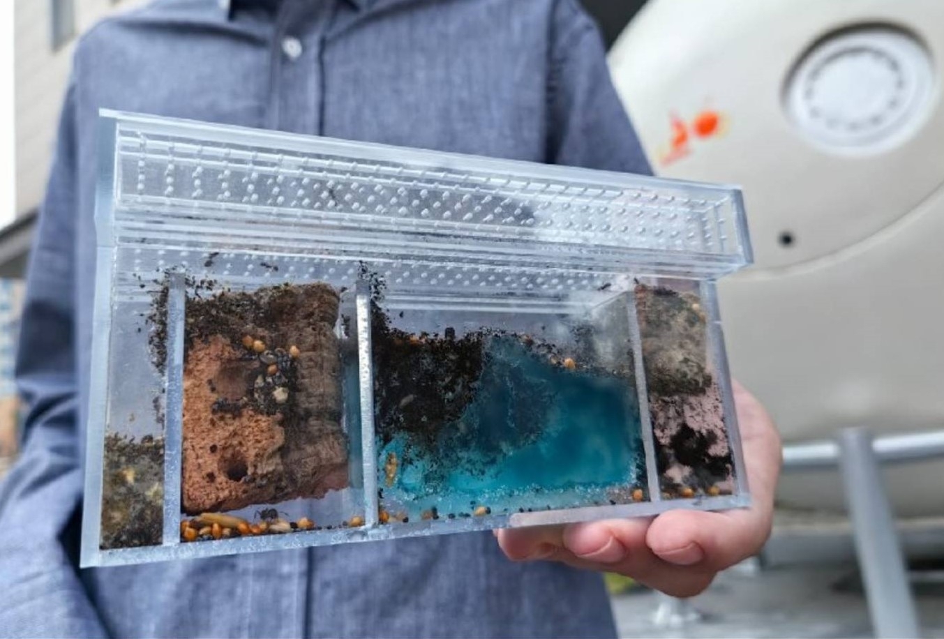 A person in a blue clean suit holds up a transparent container filled with different types of soil.