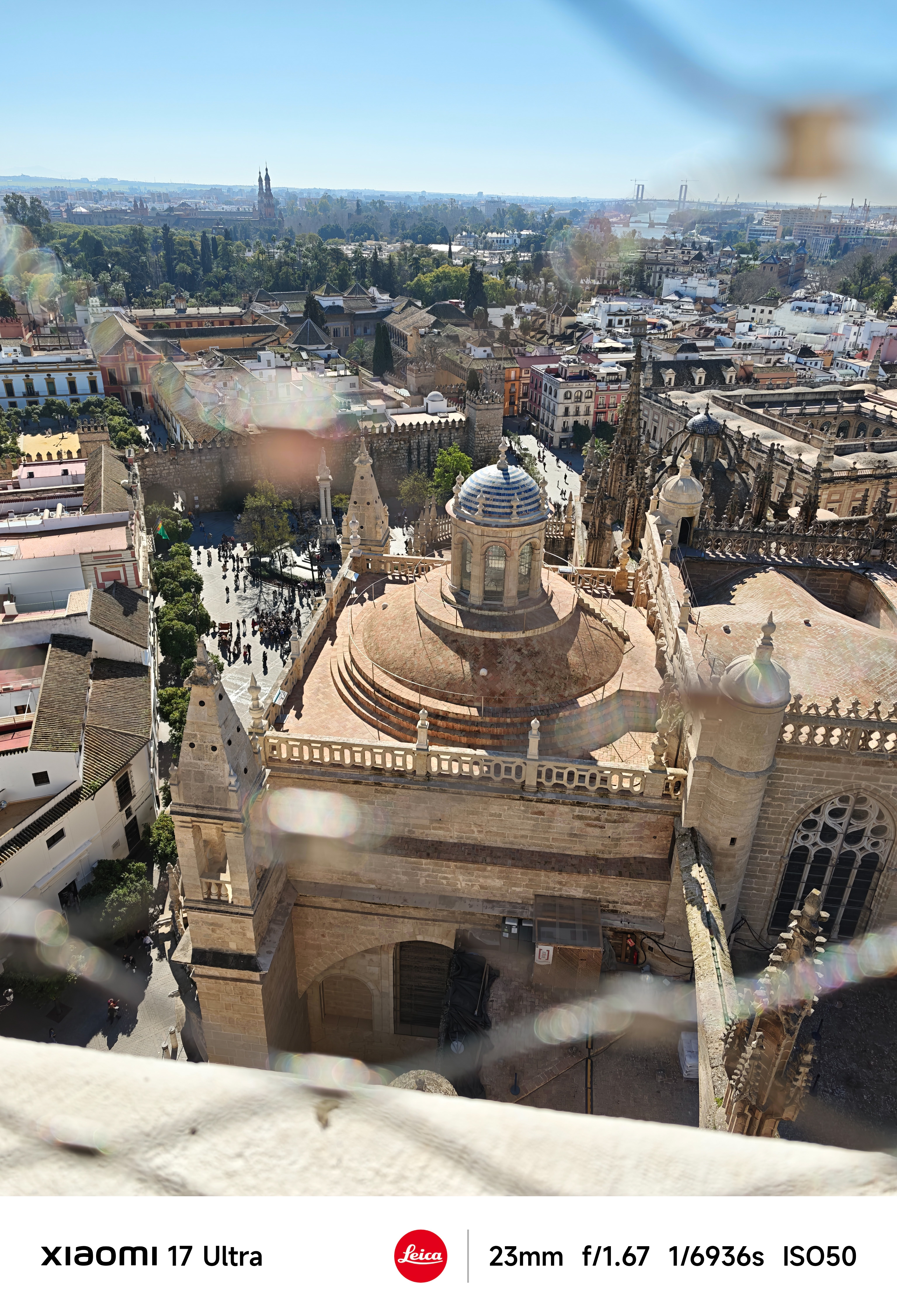 Elevated view over Seville Cathedral rooftops with lens flare and the city skyline beyond.