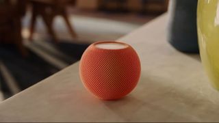 The Apple HomePod Mini in orange on a pale wooden surface.