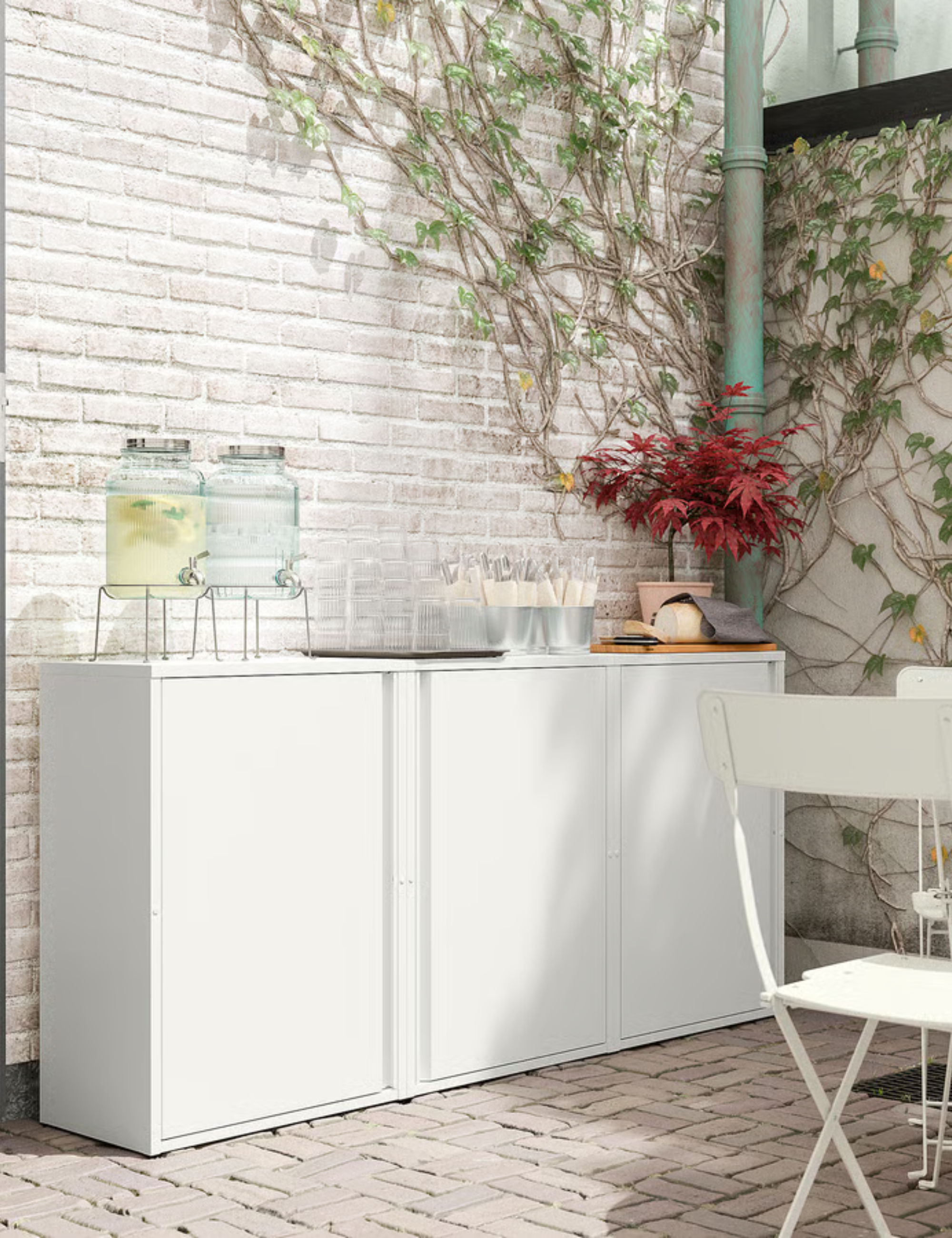Trio of white IKEA SUNDS&amp;Ouml; outdoor cabinets topped with beverages dispensers and assorted tableware in the corner of a brick patio area with crawling vines