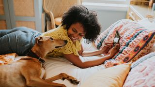 Young woman lying on bed holding phone and looking at lurcher dog beside her