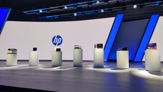 A range of HP devices set on pedestals on the keynote stage at HP Amplify 2025 in Nashville, with a large screen in the background bearing the HP logo against a white background. The devices include AI PCs, laptops, and printers.