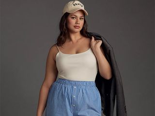 Model wears white tank tucked in boxer style pants with baseball cap while draping a leather jacket over her shoulders