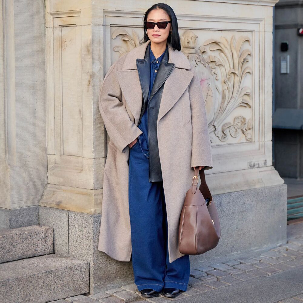 A woman is pictured wearing a blue denim set with a black leather blazer layered over it. Her outfit also includes an oversized beige coat with a large handbag and black boots. 