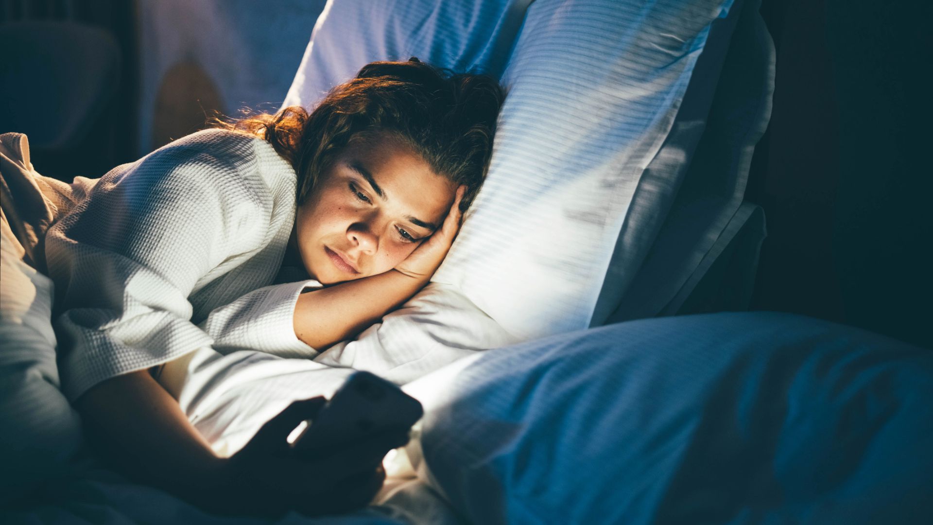 A young woman lies on her side in bed, her face illuminated by her phone screen as she 'brain rots'