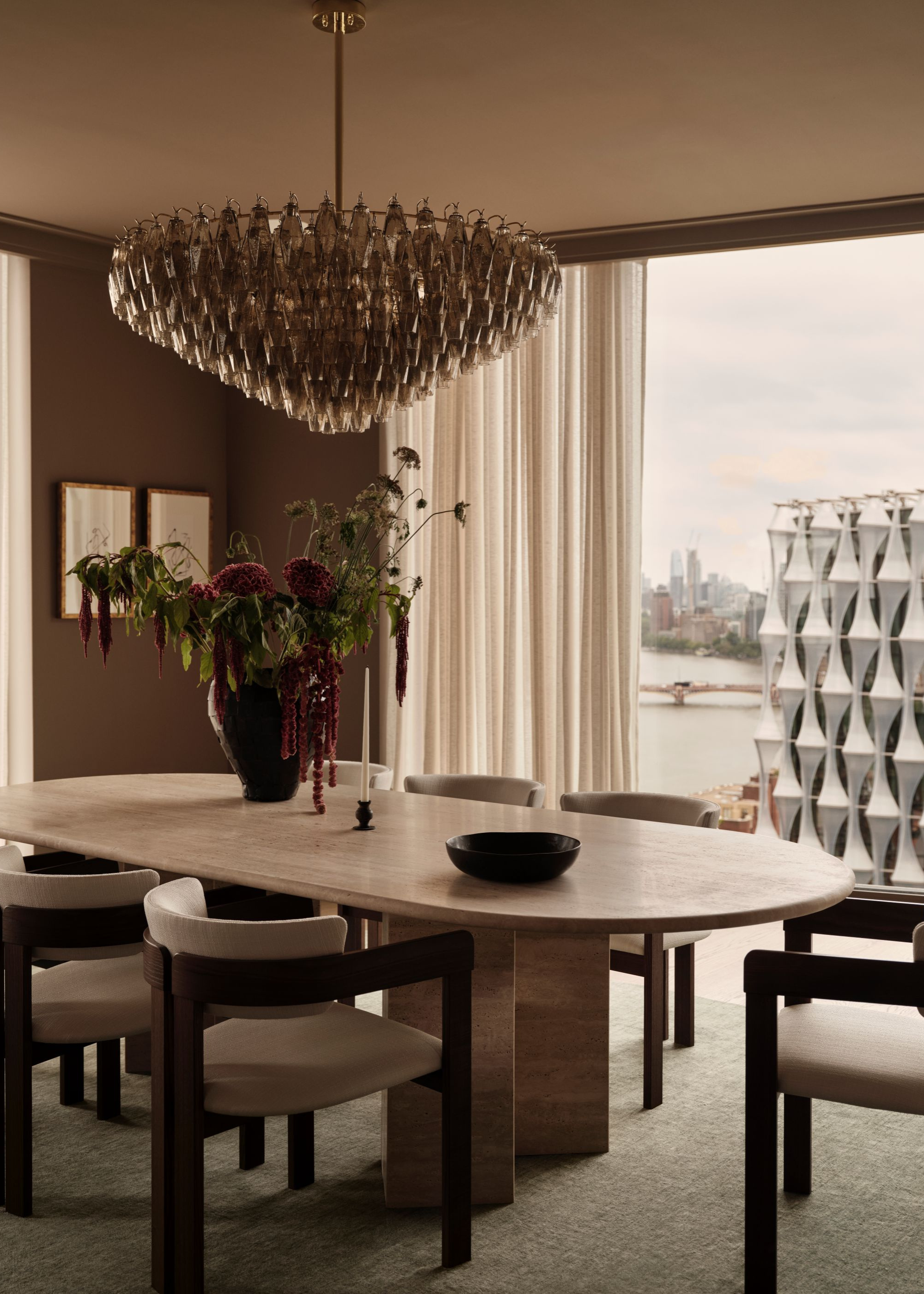 A modern dining area with a large chandelier hanging above the curved dining table with a large vase of decorative flowers and foliage on top and views of the London skyline can be seen through the full length window partly closed with sheer cream curtains