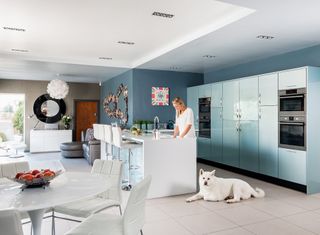 a contemporary kitchen with all white floors and island, dining table and side table, and blue cabinets, with a woman washing up at the kitchen island next to a white dog