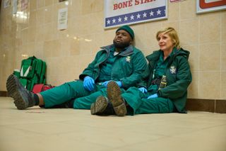 Maleek (Samson Kayo) and Wendy (Jane Horrocks) sit on the floor of a chicken shop in their paramedic uniforms with their backs to the wall - Maleek has a dejected look on his face, and Wendy looks concerned