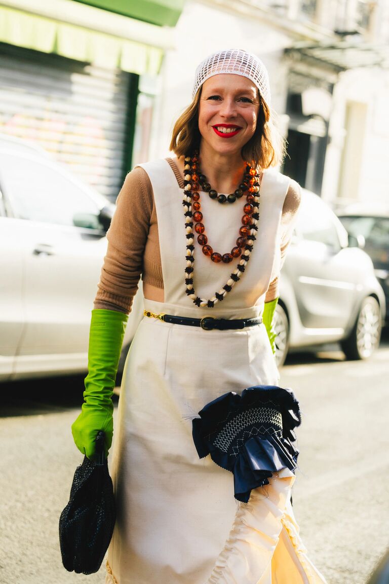 a photo of Chloe King at New York Fashion Week wearing a white dress, brown sweater, green gloves, and multicolored beaded necklaces