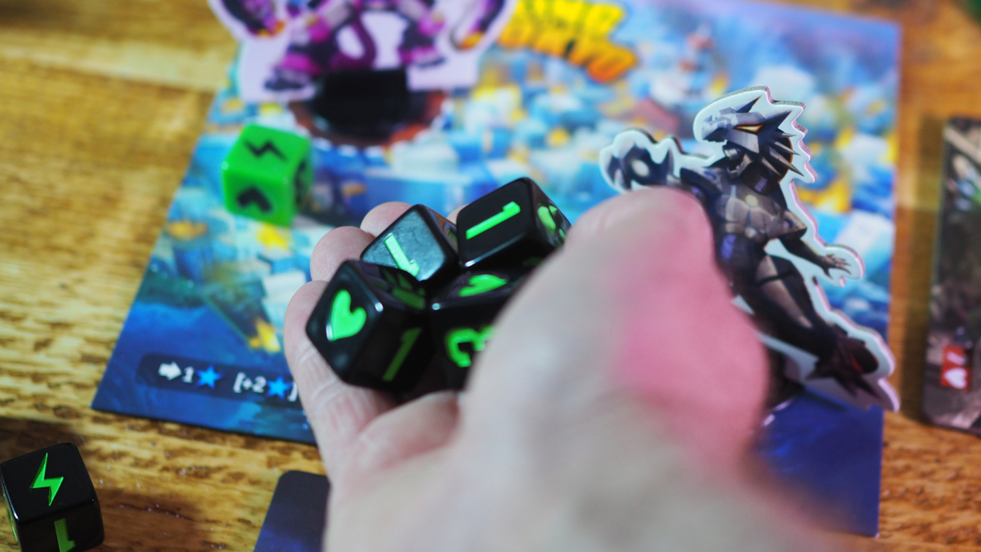 A hand holds out King of Tokyo dice, with the board and tokens in the background