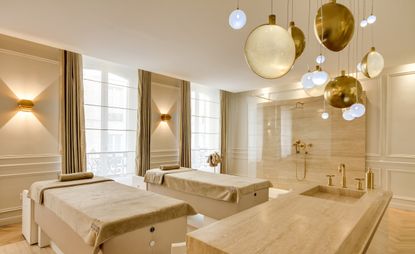 A treatment room in a spa, with two massage tables, a sink and a shower, all decorated in ivory and marble