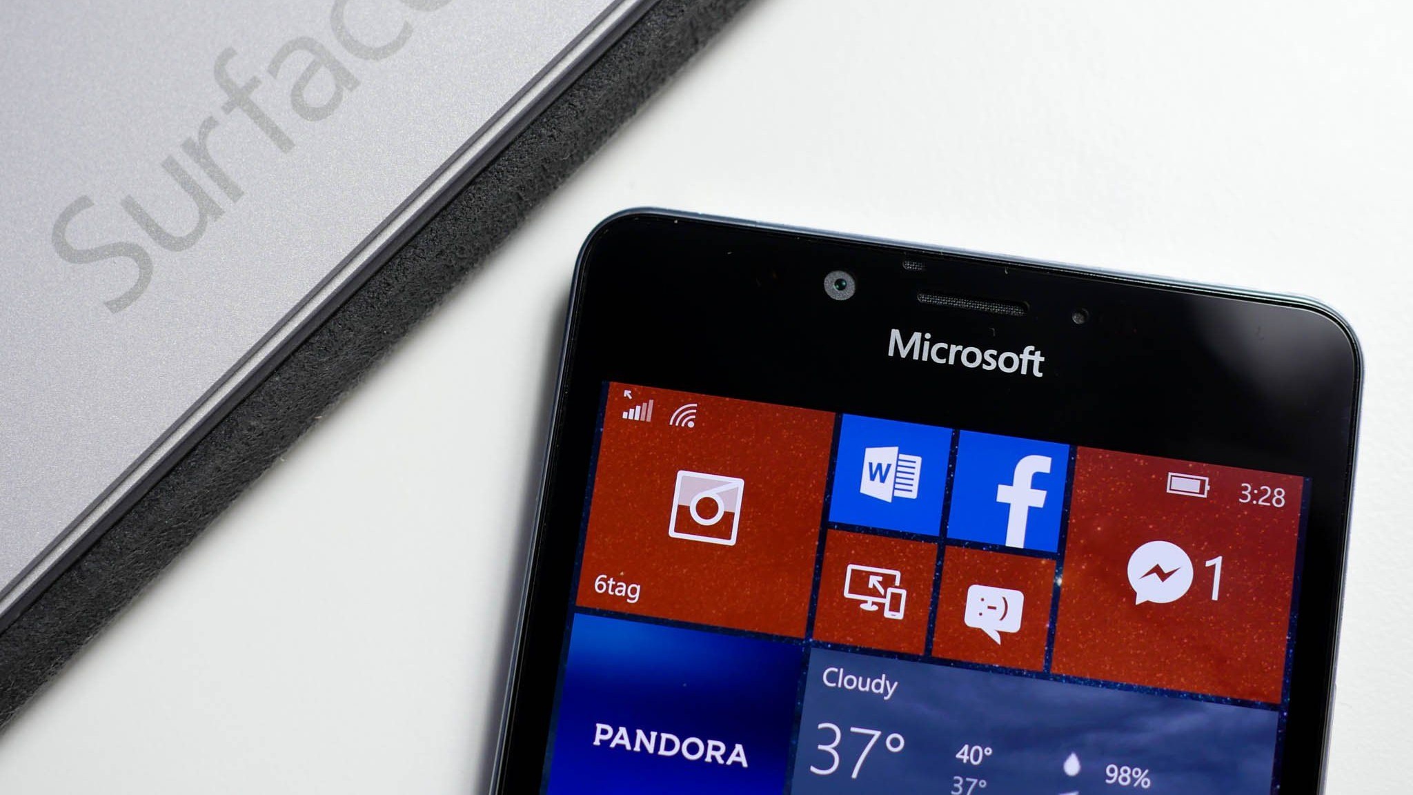 Close-up of a Microsoft smartphone displaying colorful app tiles like Word, Facebook, and Messenger. A Surface tablet is partially visible beside it.