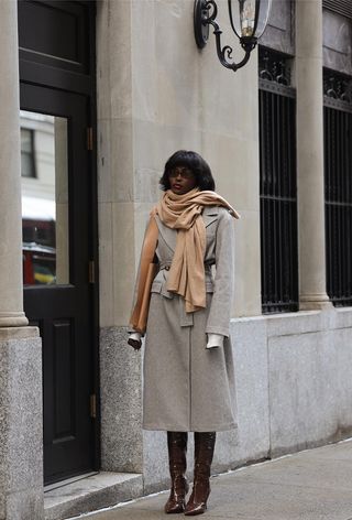 animal print boot trend is shown on a woman standing on the sidewalk wearing a tan scarf with a gray coat and brown crocodile-embossed knee-high boots