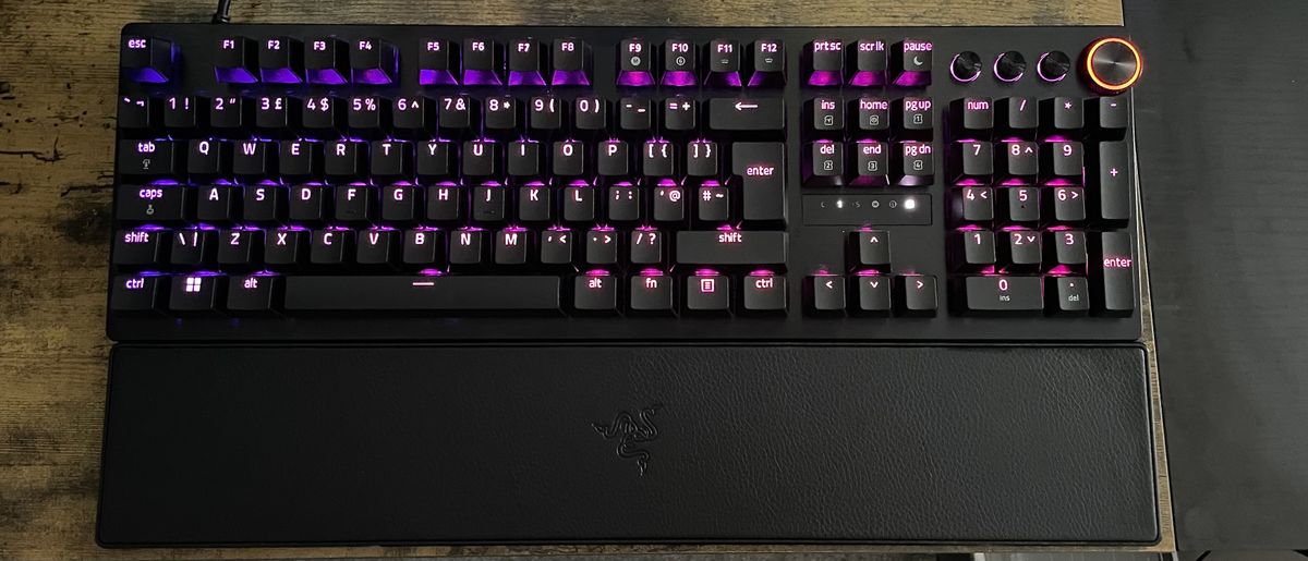 A Razer Huntsman V3 Pro shown on a wooden desk top with purple and pink backlighting behind the keys.