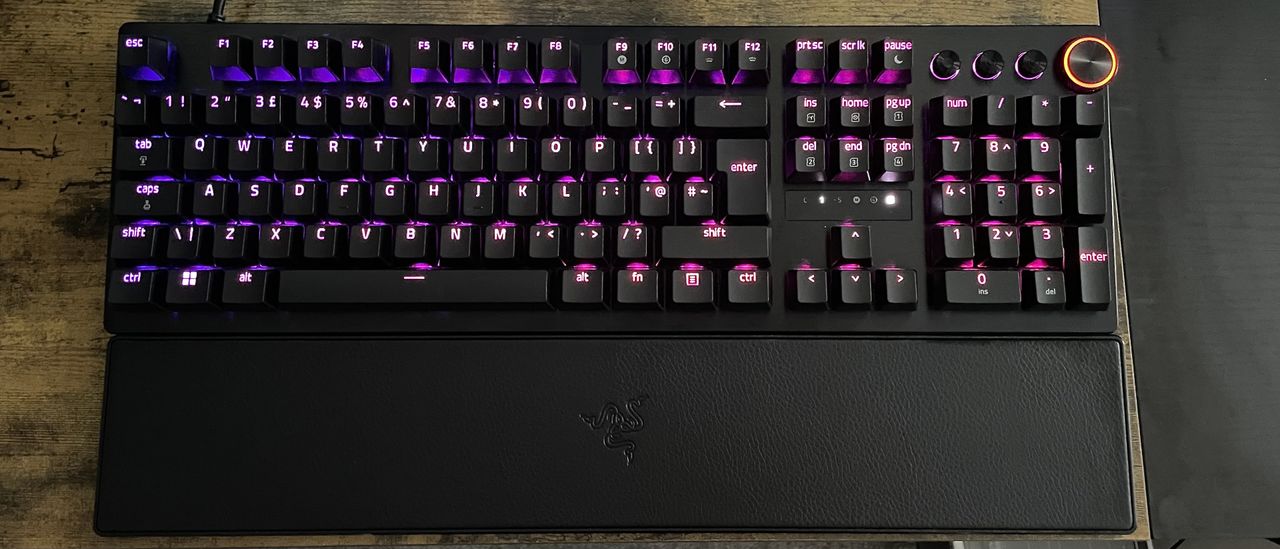 A Razer Huntsman V3 Pro shown on a wooden desk top with purple and pink backlighting behind the keys.