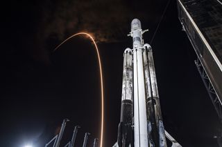 A triple-booster Falcon Heavy rocket is seen from close to its base, looking upward against the backdrop of a black sky. The rocket and four water deluge spigots, which from this perspective scale down in size the closer they are to the rocket, are lit with a bright white light, which also reveals the tall black launch tower. The rocket's core booster is a clean white, while the side boosters are covered with patterns of black soot. Between the rocket and the spigots, a thin light streak from the launch of a different rocket climbs the black sky and curves as it reaches altitude. The curve bends at the same height as the nose of Falcon Heavy.