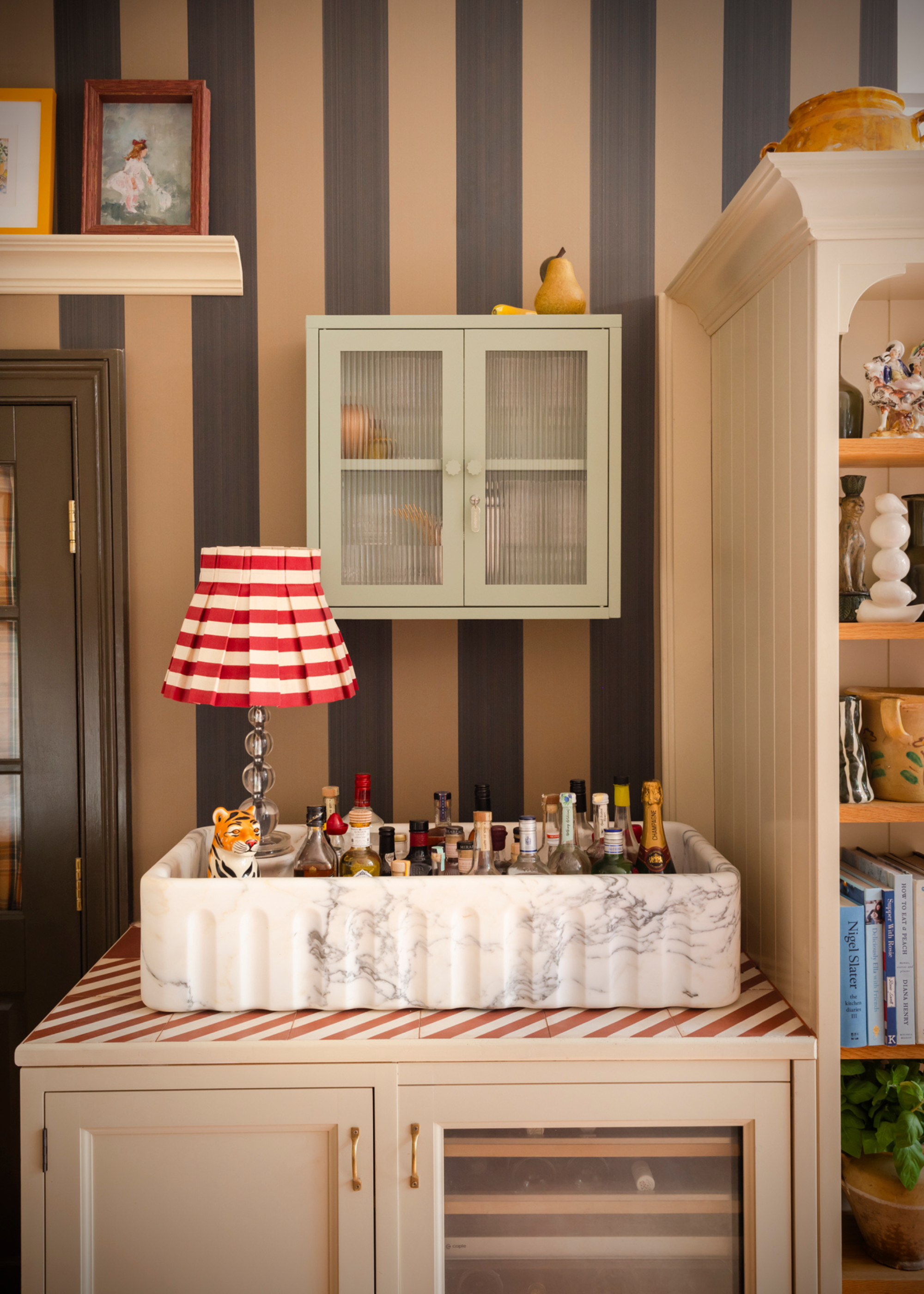 A stone sink with a collection of bottles, a wine rack below, a unit with shelves holding vases, books and trinkets. A light green fluted glass cabinet on a wall with striped wallpaper.