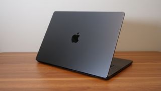 An open, dark gray laptop viewed from the back, on top of a wooden desk against a white wall.