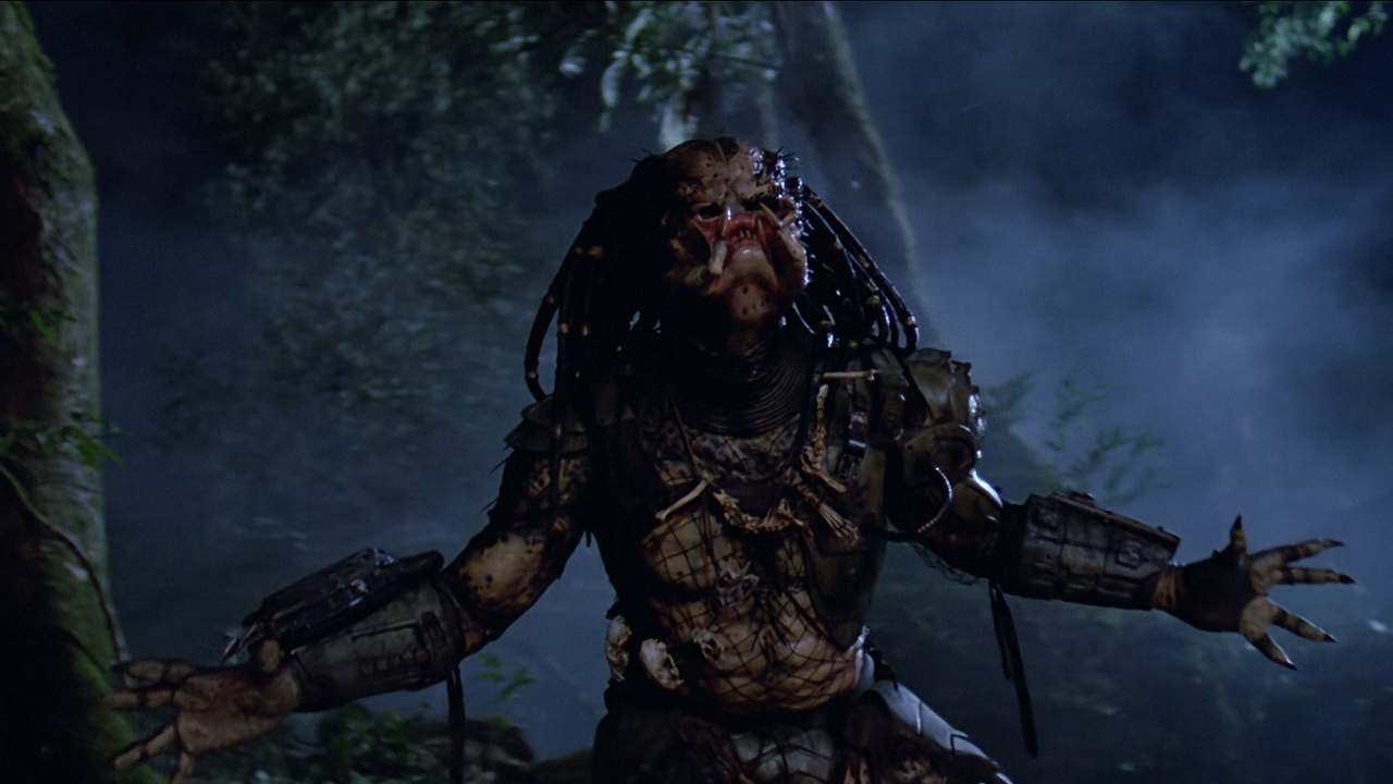 Still from the movie Predator.  Here we see Predator without most of their armor, throwing their head and arms back in a battle cry.