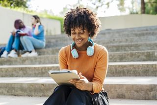 Student sitting on steps outside, looking at notepad and wearing headphones around her neck
