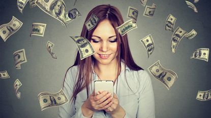 A young woman looks down at her phone while cash rains down around her.