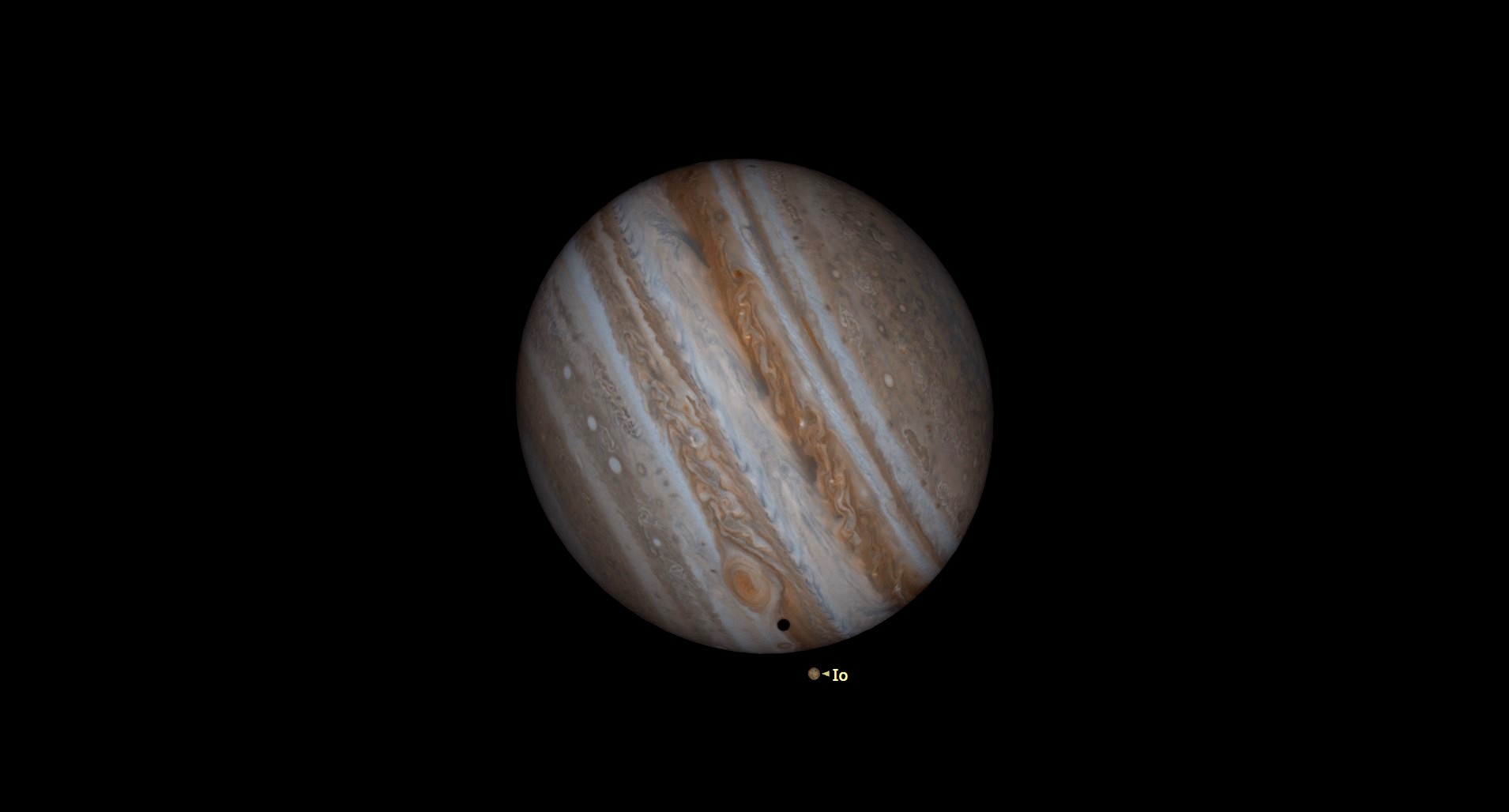 Un Jupiter mare, gazos, se înclină spre dreapta, straturile sale multicolore de maro, bronz și portocaliu atârnând în spațiul negru.  aproape de fundul planetei, furtuna cu ochi roșii a lui Jupiter se învârte deasupra umbrei negre aruncate pe suprafața planetei de pe luna Io, odihnindu-se chiar dedesubt și etichetată.