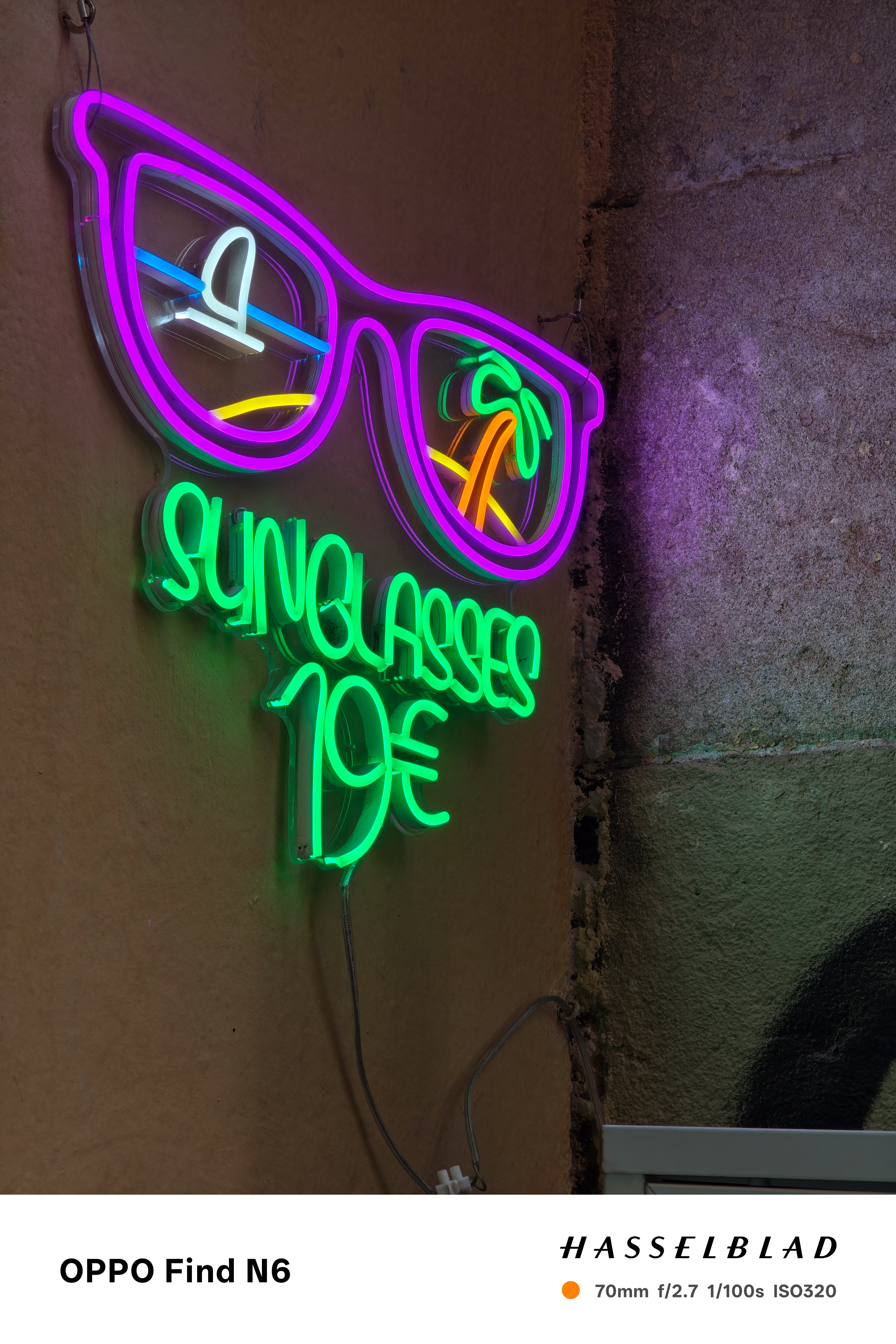 A vertical shot of a neon sign mounted on a textured tan wall. The sign is shaped like a large pair of purple sunglasses with green palm trees inside the lenses, with the words "SUNGLASSES 19&euro;" glowing in bright green neon below.