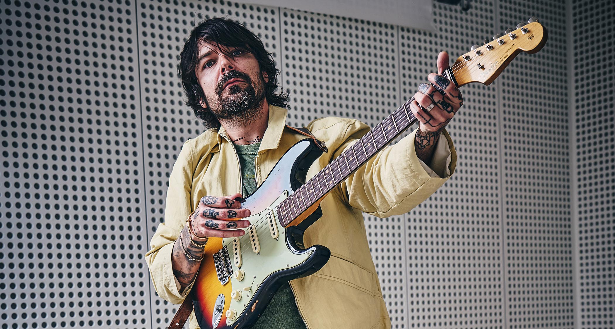 Simon Neil of Biffy Clyro wears a custard-colored jacket and green T-shirt and poses with a sunburst Fender Stratocaster