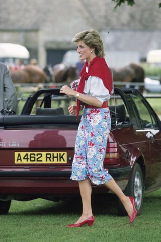 Princess Diana wearing a blue floral skirt with red heels, a white top and a red sweater around her neck walking in grass in front of a maroon car