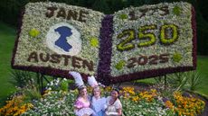 Three women dressed in Regency outfits stand in front of a floral decoration honoring Jane Austen
