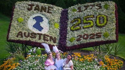 Three women dressed in Regency outfits stand in front of a floral decoration honoring Jane Austen