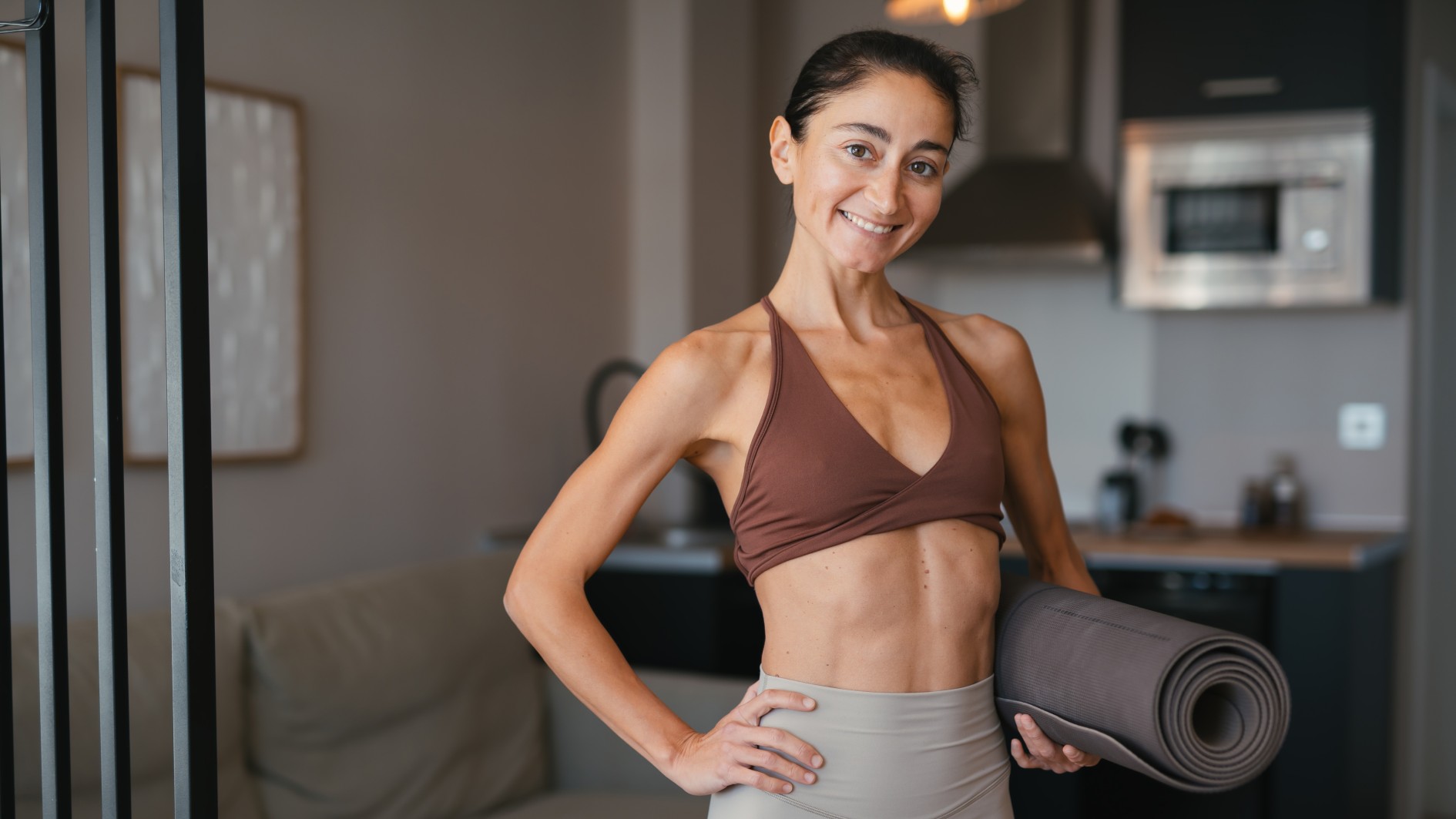 Fit woman in activewear holding yoga mat at home, smiling to camera, hand on hip