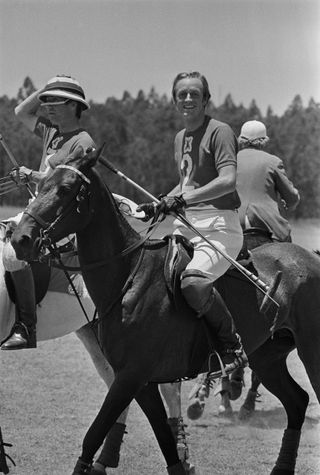 A black and white photo of Andrew Parker Bowles riding a horse in a tee shirt and white pants