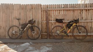 Two loaded gravel bikes stand in front of a wooden fence in the shade