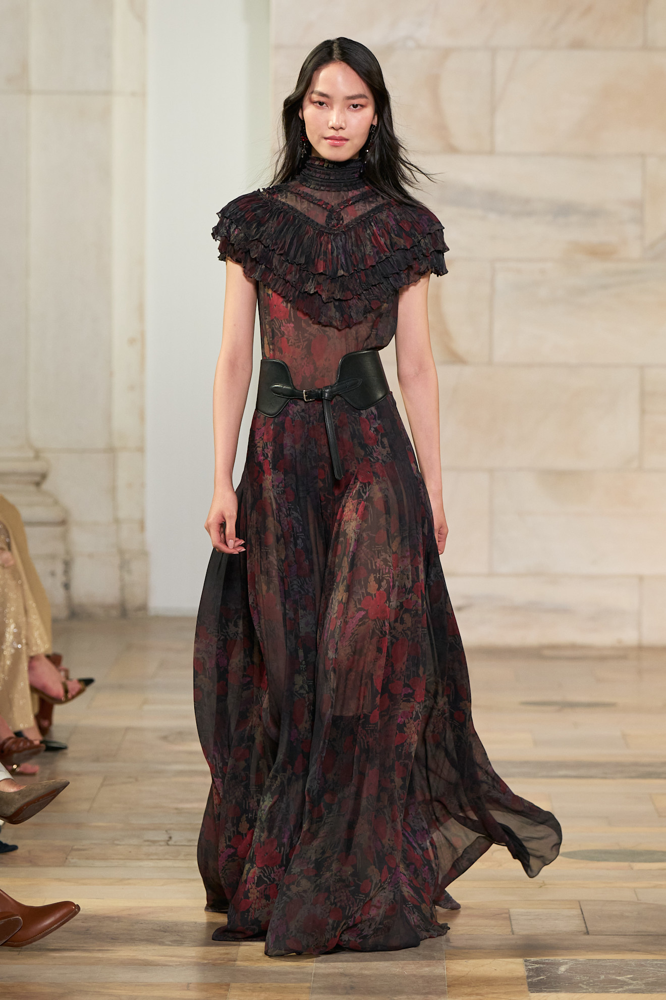 Model walking in the Ralph Lauren Fall Winter 2025 runway show wearing a black and red boho, sheer maxi dress with shoulder ruffles, plus a large leather waist belt.