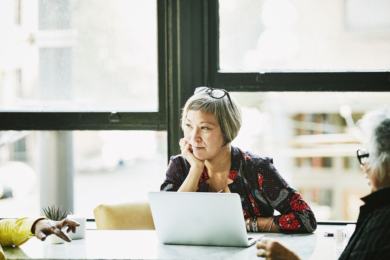 An older woman with an open laptop sits in a meeting.