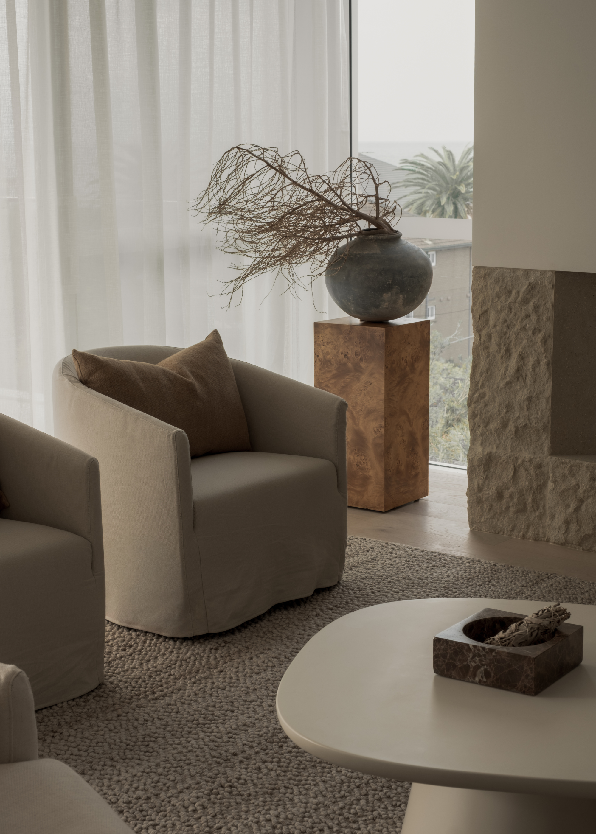 A muted minimalist living room with beige sofa seats, a textured rug, a burl wood plinth with a vase and stems, a textured fireplace, a small stone bowl with dried herbs, and sheer window treatments