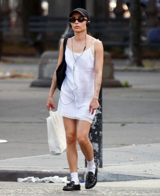 a street style image of Zoë Kravitz wearing a white mini dress, black baseball hat, black loafers, and white crew socks, and listening to music with over-the-head headphones