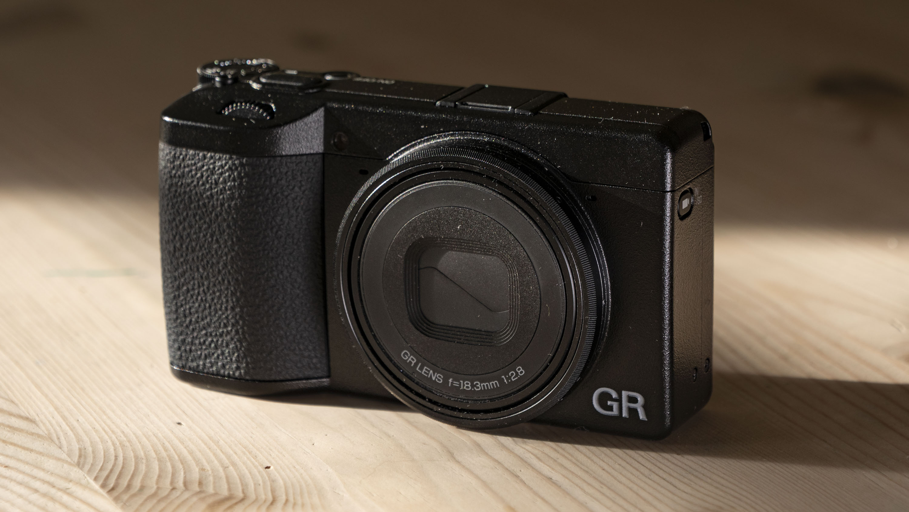 Ricoh GR IV compact camera on a wooden table, with strong sidelight and dark shadows