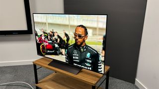 The 48-inch LG C4 OLED TV on a TV stand at an angle. On screen is F1 driver Lewis Hamilton in his racing overalls with an F1 car in the background.