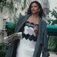 model wearls white slip dress with lace details and a long gray trench coat while holding a newspaper and walking 