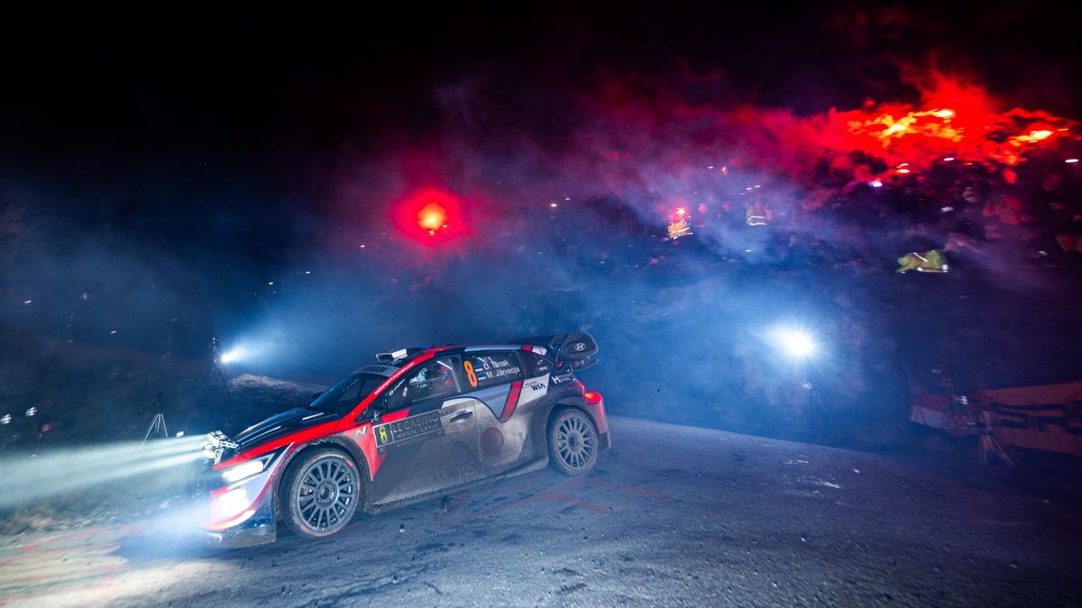 A rally car drives through the night, illuminated by headlights and flares at the Monte Carlo rally