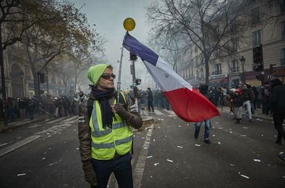 PARIS, FRANCE - DECEMBER 05: A Gilet Jaune, or Yellow Vest' holds a French Tricolor amidst tear gas as protestors and French Riot Police clash during a rally near Place de Republique in suppo