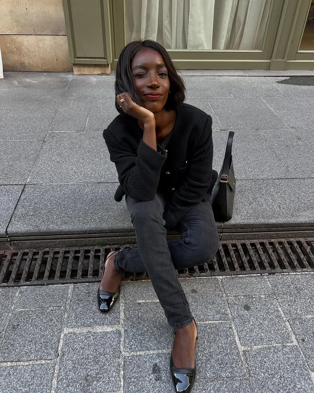 @aidabadji_ wears black skinny jeans with a boucle jacket and ballet flats while sitting on a pavement 