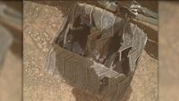 a close-up of a metal wheel with deep cracks and caved-in sections as it rests on a reddish-orange rocky surface