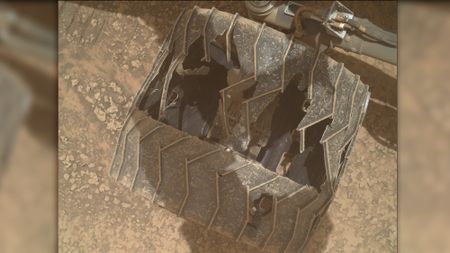 a close-up of a metal wheel with deep cracks and caved-in sections as it rests on a reddish-orange rocky surface