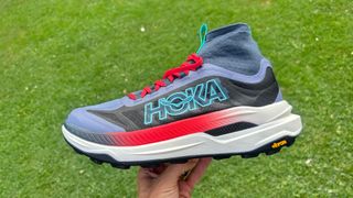 Person holding the Hoka Tecton X3 trail running shoes in front of a grass field