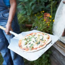 a woman taking out a pizza from a homemade pizza oven - GettyImages-1043643456